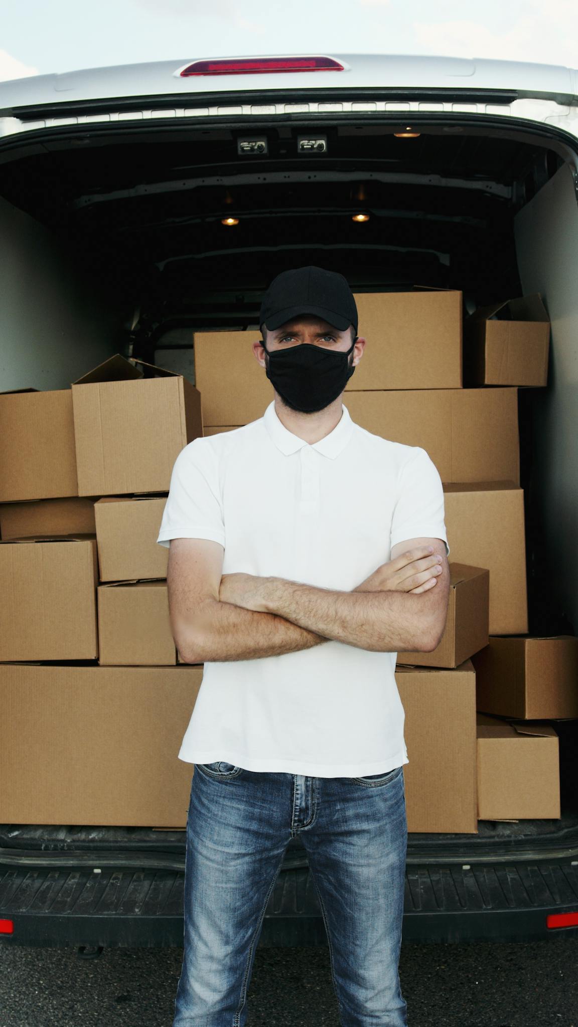 Home A courier wearing a mask standing in front of a loaded van with cardboard boxes, showcasing logistics.