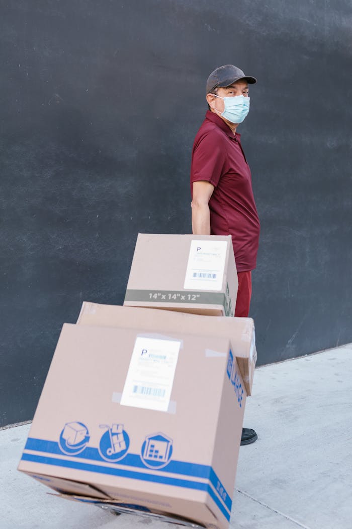 Deliveryman wearing face mask pulls cart with boxes outdoors, reflecting pandemic logistics.